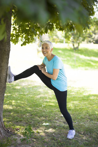 Mujer mayor hace ejercicio de estiramiento en el parque por árbol
 - Foto, imagen