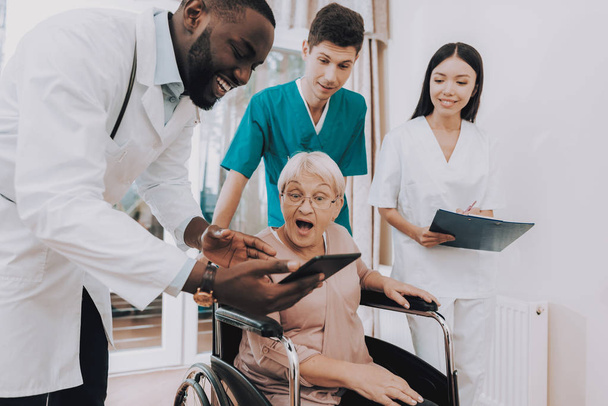 Doctor Show. Very Surprised. Elderly Woman Looks. Hospital Case. Doctor with Tablet. Patient. Patient on a Wheelchair. Nursing Home. Doctor in Clinic. Doctor Helps. Rehabilitation. Sitting Woman. - Foto, afbeelding