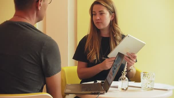 Una mujer de negocios y un hombre en su descanso con su tableta se sientan en un café con paredes amarillas y discuten un proyecto
 - Metraje, vídeo