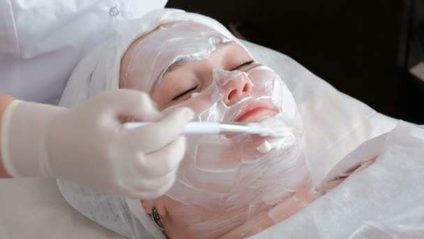 Una mujer gorda recibe un procedimiento de limpieza y elevación de la piel de la cara en la oficina de salones de belleza. Las manos del cosmetólogo en guantes untan con un cepillo una crema blanca en la frente de una niña
 - Metraje, vídeo