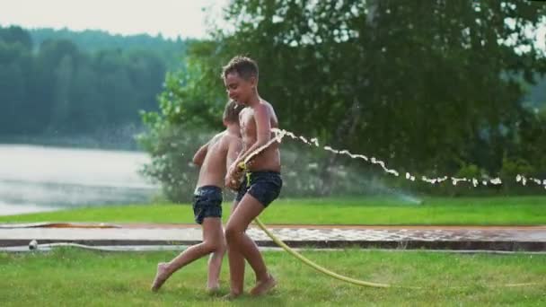 Junge in Badehose gießt Wasser auf seinen jüngeren Bruder, der sich im Park auf der Wiese am See vergnügt - Filmmaterial, Video