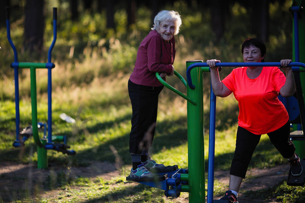Mujer mayor en el patio de recreo deportivo haciendo ejercicios en el simulador con el entrenador
. - Foto, imagen