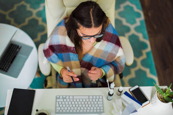 Вид зверху на молоду хвору жінку, яка сидить в офісі і тримає ручку в руці і дивиться на ліки
 - Фото, зображення