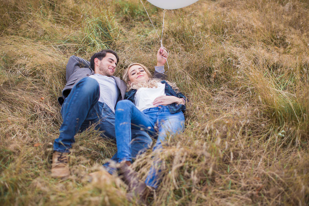 The young happy romantic couple lying in embrace on the grass outdoor and boyfriend holding the white balloons in hand - Фото, изображение