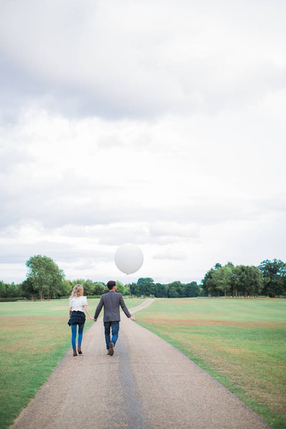 Back view of the young happy romantic couple in love who walking outdoor and boyfriend holding balloon in hand - Fotó, kép