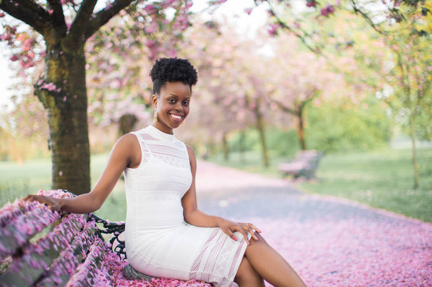 Portrait of the young happy pretty woman who sitting on the bench with pink petals in the park and smiling - Photo, Image