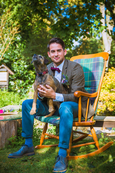 El joven guapo y sonriente prometido sentado en la silla de madera con su perro en el jardín
 - Foto, imagen