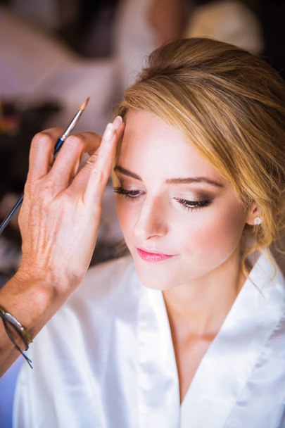 The makeup artist putting makeup on face of the young beautiful bride indoors - Valokuva, kuva