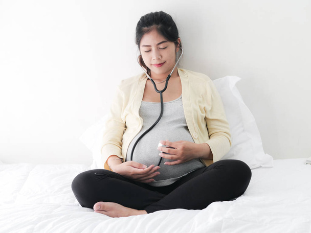 Asian pragnant woman using stethoscope listening her baby on bed, lifestyle concept. - Фото, зображення