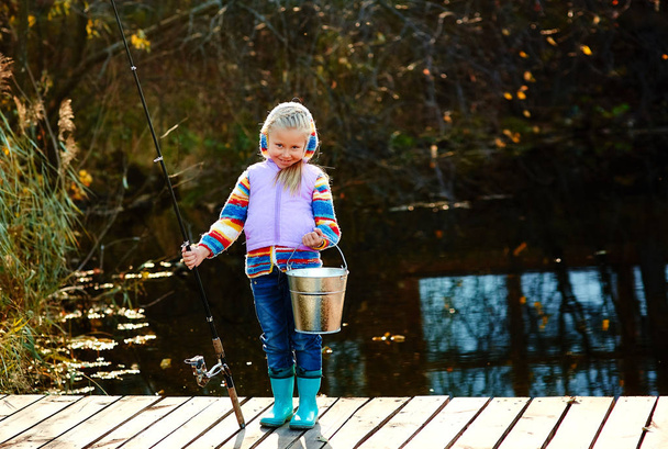 An angler girl with a fishing rod and a bucket with a catch stands on the bridge. - Zdjęcie, obraz