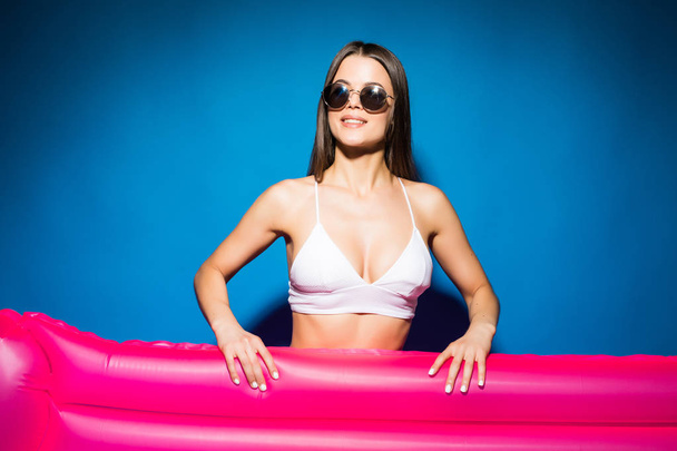 Hermosa mujer en gafas de sol con bikini blanco sosteniendo con colchón rosa aislado sobre fondo azul
 - Foto, imagen