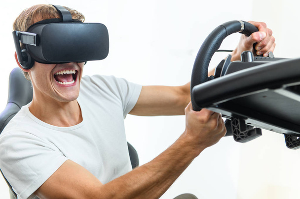 A young, handsome guy in a white T-shirt, playing in a race, on a simulator of virtual reality - Photo, Image