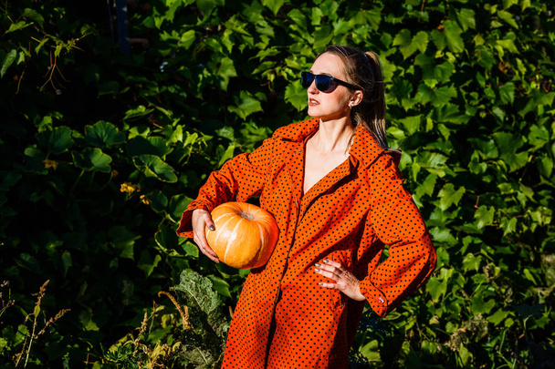 a young white woman in the orange coat on a background of green leaves with a pumpkin in the hands - Foto, afbeelding