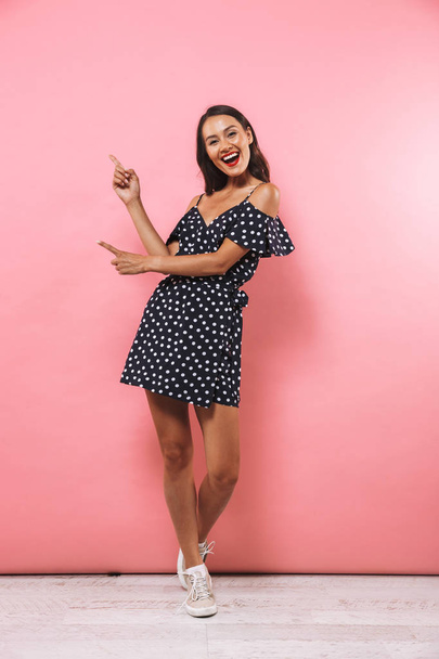 Full length image of Cheerful brunette woman in dress pointing on copyspace and looking at the camera over pink background - Фото, изображение