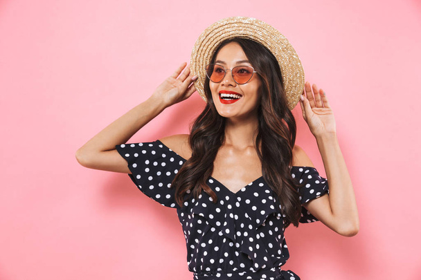 Задоволена брюнетка в солом'яному капелюсі і сонцезахисних окулярах позує, дивлячись на рожевий фон
 - Фото, зображення