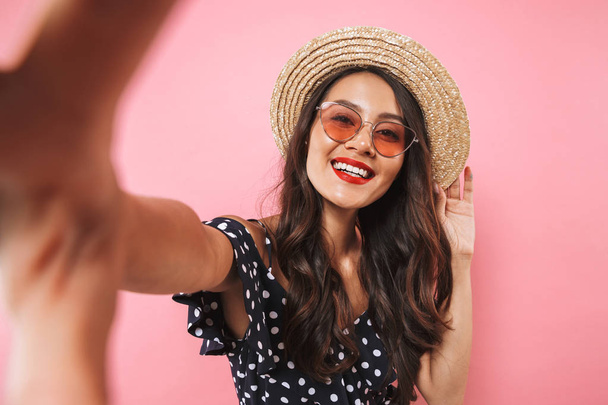 Ευτυχισμένη γυναίκα μελαχρινή σε ψάθινο καπέλο και γυαλιά ηλίου κάνοντας selfie ενώ κοιτάζοντας την κάμερα σε ροζ φόντο - Φωτογραφία, εικόνα