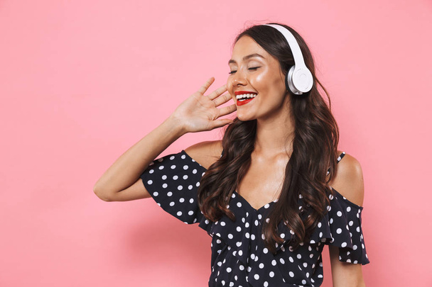 Lachende brunette vrouw in jurk en koptelefoon luisteren van muziek met gesloten ogen op roze achtergrond - Foto, afbeelding