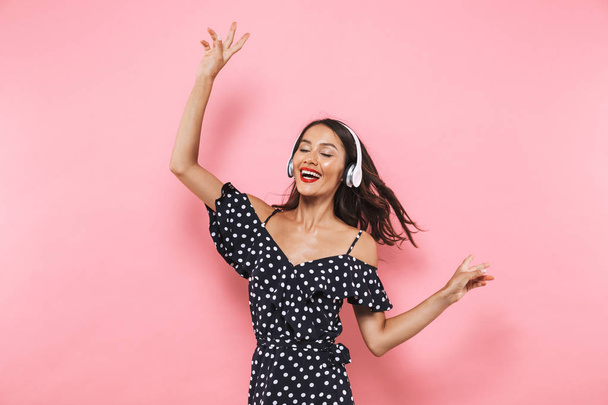 Pleased brunette woman in dress and headphones listening music with closed eyes while showing peace gestures and enjoys over pink background - Fotografie, Obrázek