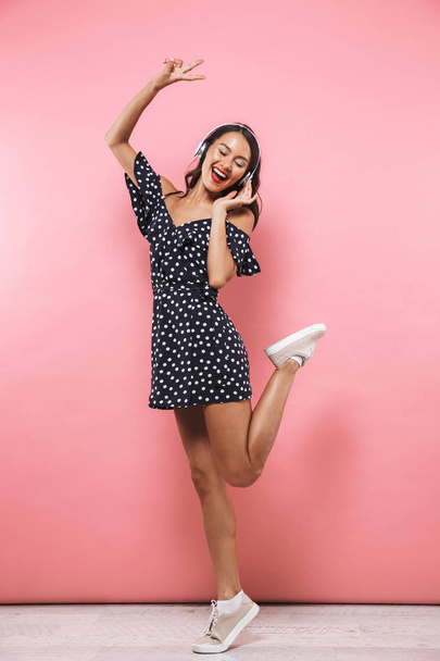 Full length image of Joyful brunette woman in dress and headphones listening music while jumping and enjoys over pink background - Fotografie, Obrázek