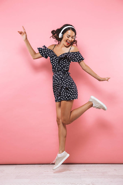Full length image of Cheerful brunette woman in dress and headphones listening music while jumping over pink background - Fotoğraf, Görsel