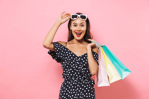 Surprised happy brunette woman in dress take off sunglasses and looking at the camera while holding packages over pink background - Fotografie, Obrázek