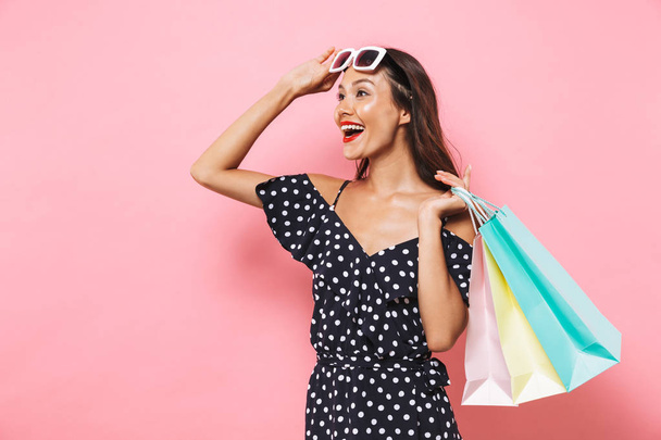 Surprised happy brunette woman in dress and sunglasses holding packages while looking away over pink background - Zdjęcie, obraz