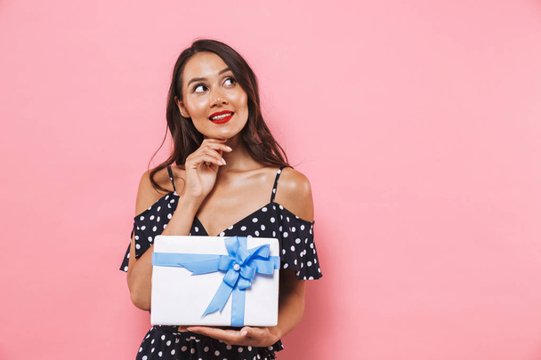 Photo of happy thinking woman isolated over pink background holding susprise gift box. - Zdjęcie, obraz