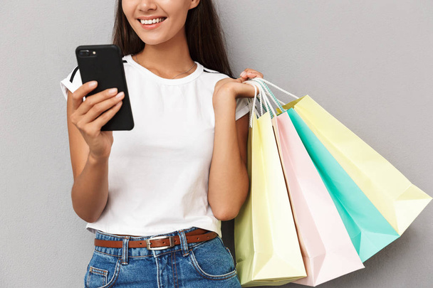 Cropped image of young cheerful woman holding shopping bags isolated over grey background using mobile phone. - Foto, immagini
