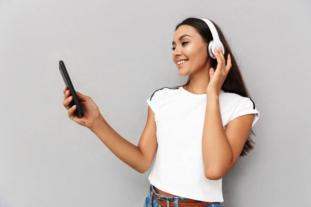 Image of young happy emotional woman using mobile phone listening music with headphones isolated over grey background. - Fotoğraf, Görsel