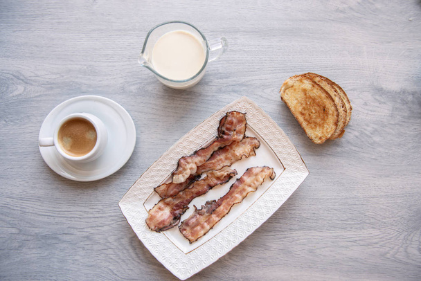 Gebakken spek op wit bord met kopje koffie en melk kruik - Foto, afbeelding