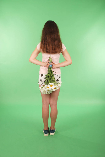 Widok z tyłu młoda dziewczyna z bukiet rumianku w ręce patrząc na ścianie. Na białym tle na zielonym tle - Zdjęcie, obraz