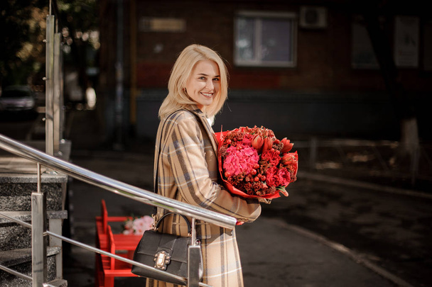 Mujer rubia feliz y sonriente en abrigo a cuadros con un gran ramo rojo de flores en el día de otoño
 - Foto, imagen
