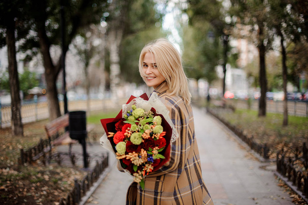 Smiling blonde woman in plaid coat holding a bright bouquet of flowers walking on alley in the autumn park - Fotoğraf, Görsel