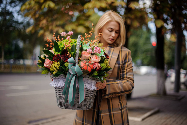 Young blonde woman holding a big wicker basket of autumn flowers against the background of city - Foto, Bild