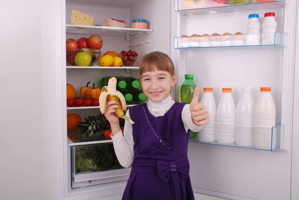 La chica sostiene un plátano en el fondo del refrigerador. Hermosa chica cerca del refrigerador con comida saludable. Frutas y verduras en el refrigerador
. - Foto, Imagen