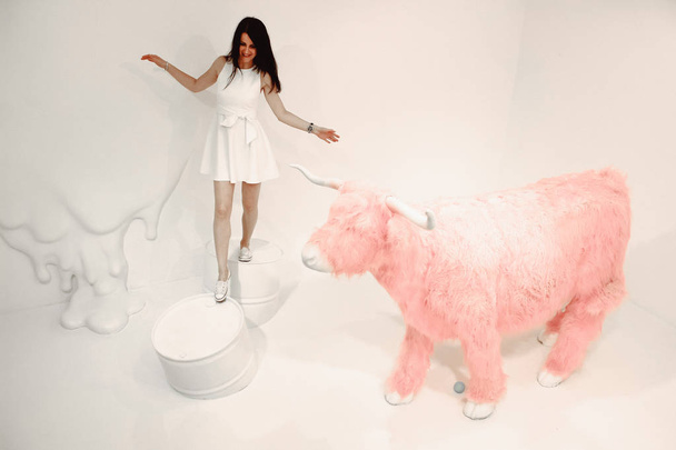Young beautiful brunette in white dress on a white background with fantastic scenery with a pink bull. - 写真・画像