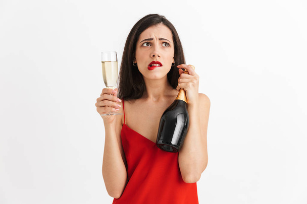 Portrait of a confused young woman in dress holding glass and bottle of champagne isolated over white background - Foto, afbeelding