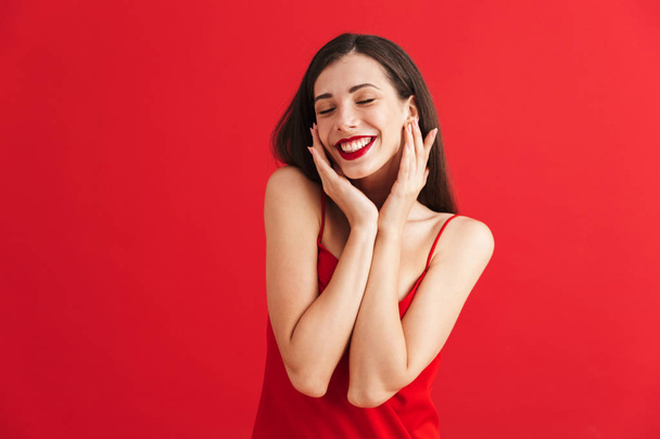 Portrait of a happy young woman in dress isolated over red background, holding hands at her face - Фото, зображення