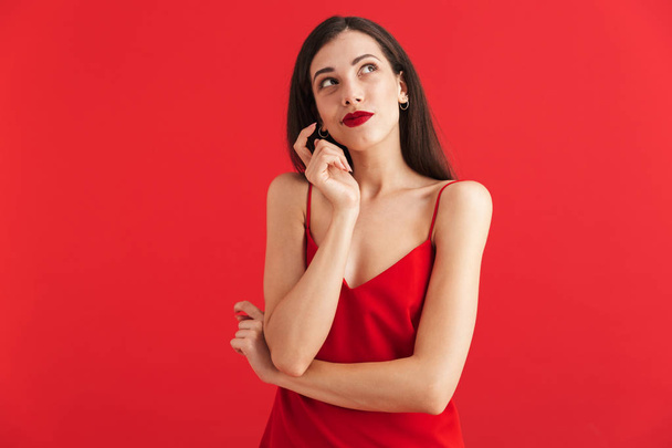 Portrait of a thoughtful young woman in dress isolated over red background, looking away - Photo, Image