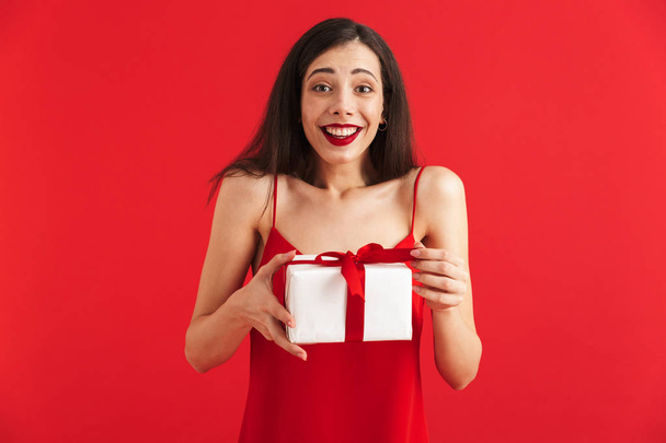 Portrait of a delighted young woman in dress holding present box isolated over red background - Foto, Imagen