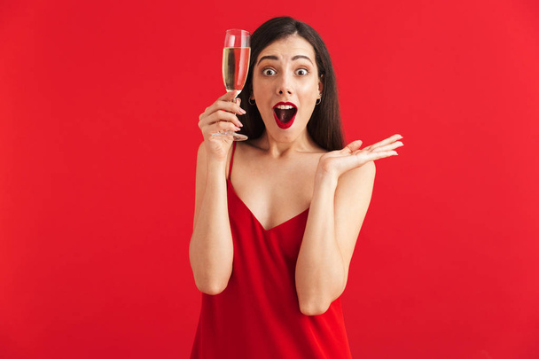 Portrait of an excited young woman in dress holding glass of champagne isolated over red background - Fotoğraf, Görsel