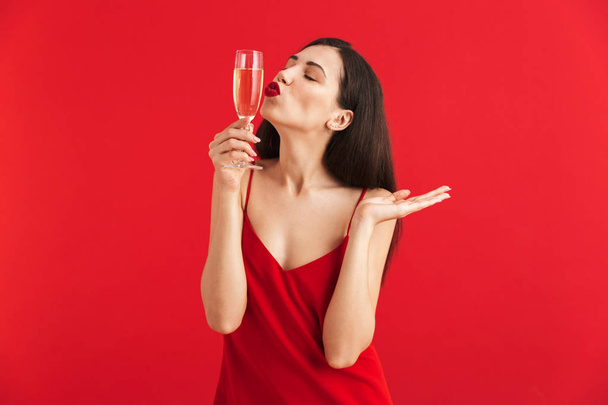 Portrait of a lovely young woman in dress holding glass of champagne isolated over red background - Fotó, kép