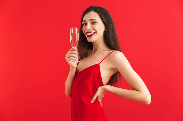 Portrait of a confident young woman in dress holding glass of champagne isolated over red background - 写真・画像