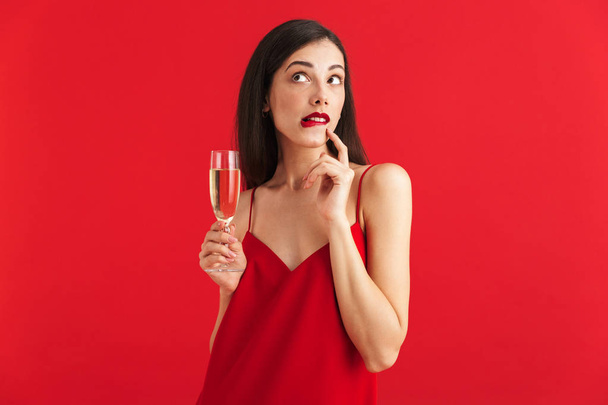 Portrait of a pensive young woman in dress holding glass of champagne isolated over red background - Valokuva, kuva