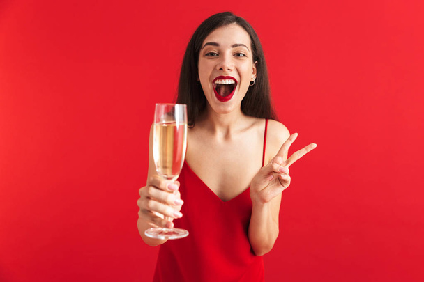 Portrait of a cheerful young woman in dress holding glass of champagne isolated over red background, showing peace - 写真・画像