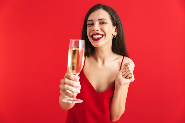 Portrait of a cheerful young woman in dress holding glass of champagne isolated over red background, toasting camera - Fotoğraf, Görsel