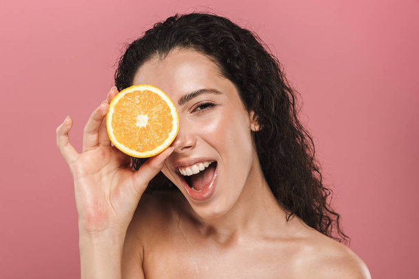 Beauty portrait of a lovely young topless woman with curly brunette hair posing over pink background, holding half of an orange - Фото, изображение
