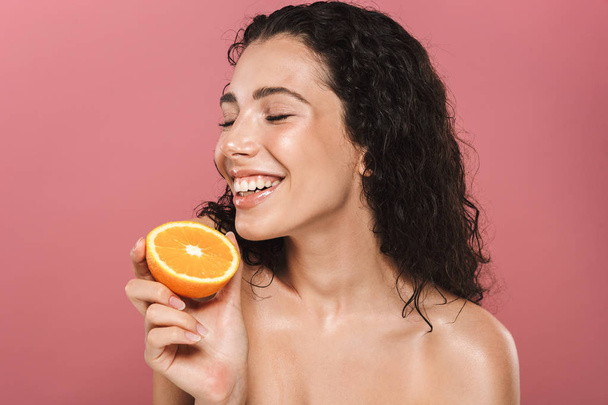 Beauty portrait of a smilng young topless woman with curly brunette hair posing over pink background, holding half of an orange - 写真・画像