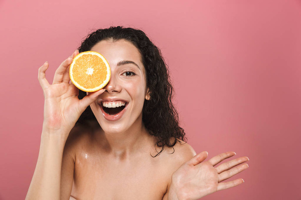 Beauty portrait of an excited young topless woman with curly brunette hair posing over pink background, holding half of an orange - Fotó, kép