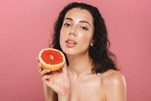 Beauty photo of woman with clean face and long hair smiling and holding piece of grapefruit isolated over pink background - Foto, Bild